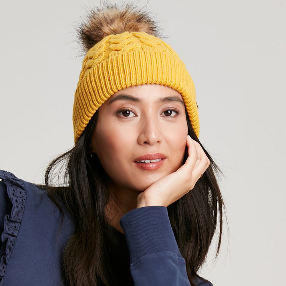 Gorro con pompón Elena punto de cable Pompón de piel sintética de Joules - Mostaza